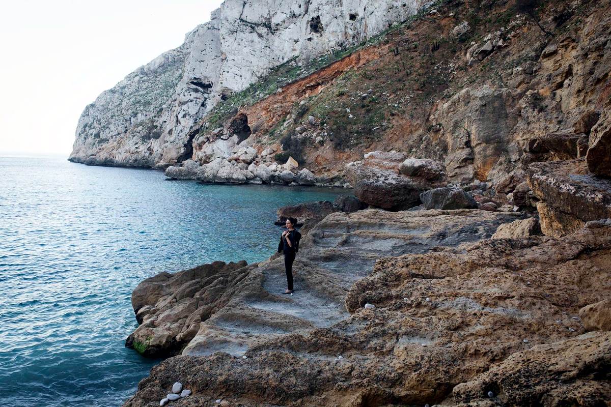A lone figure on the rocks of a small cove beneath sheer limestone cliffs